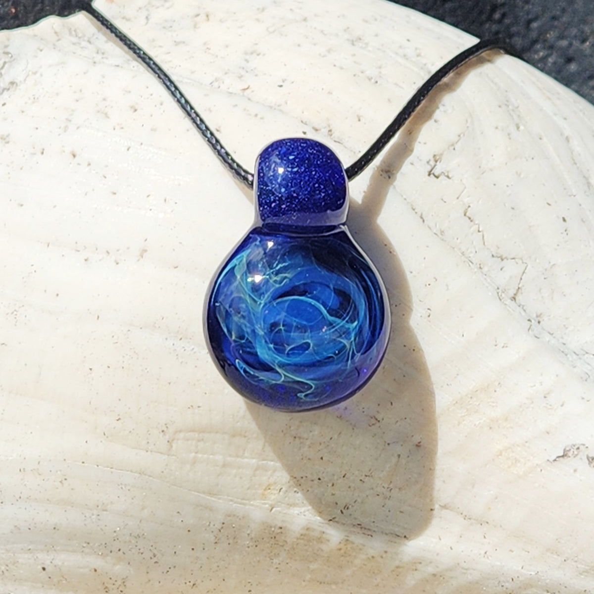 A blue glass pendant necklace with a swirling light blue pattern inside, suspended on a black cord, resting on a textured white stone surface.