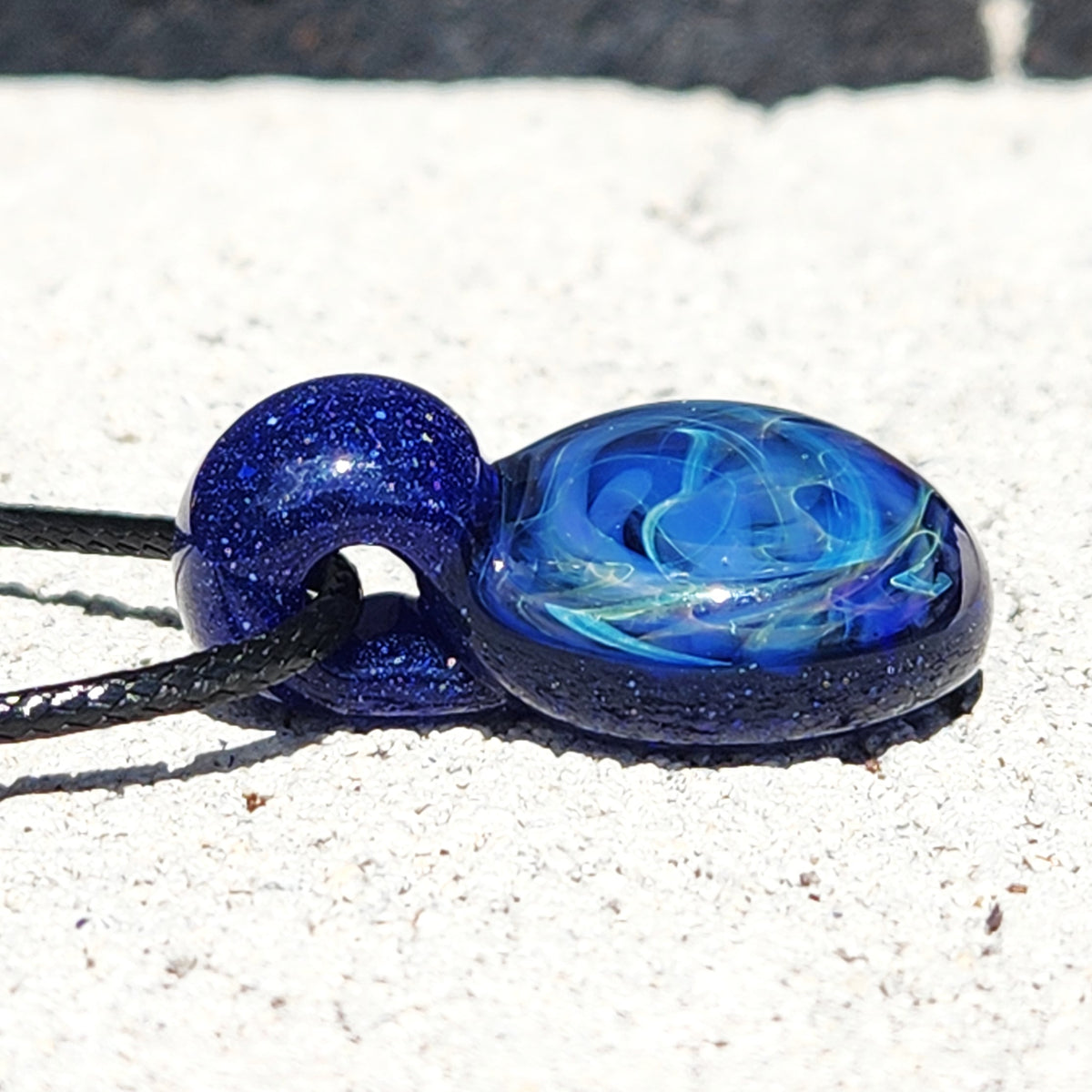 A blue glass pendant necklace with swirling light blue patterns and dark blue glittery edges, threaded on a black cord, resting on a white textured surface.
