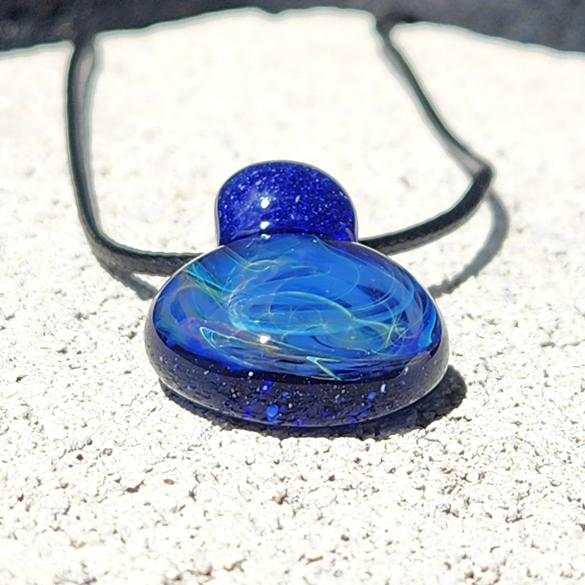 A blue glass pendant necklace with swirling light blue and white patterns and dark blue glitter, resting on a textured white stone surface with a black cord.
