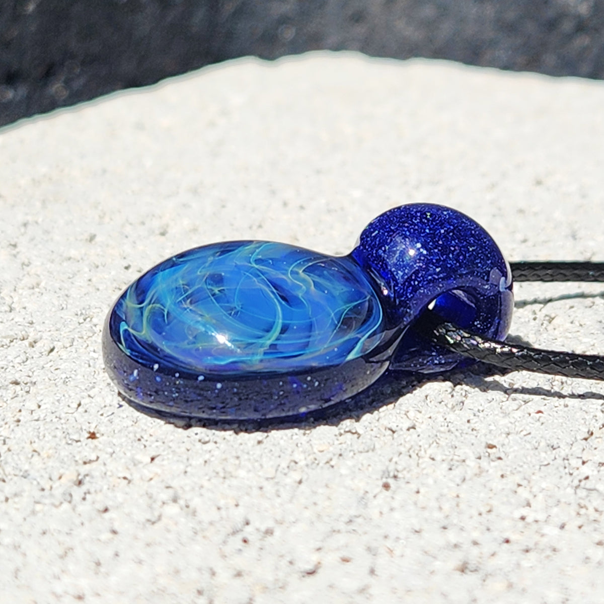 A blue and light blue marbled glass pendant necklace with a black cord, resting on a textured white stone surface.