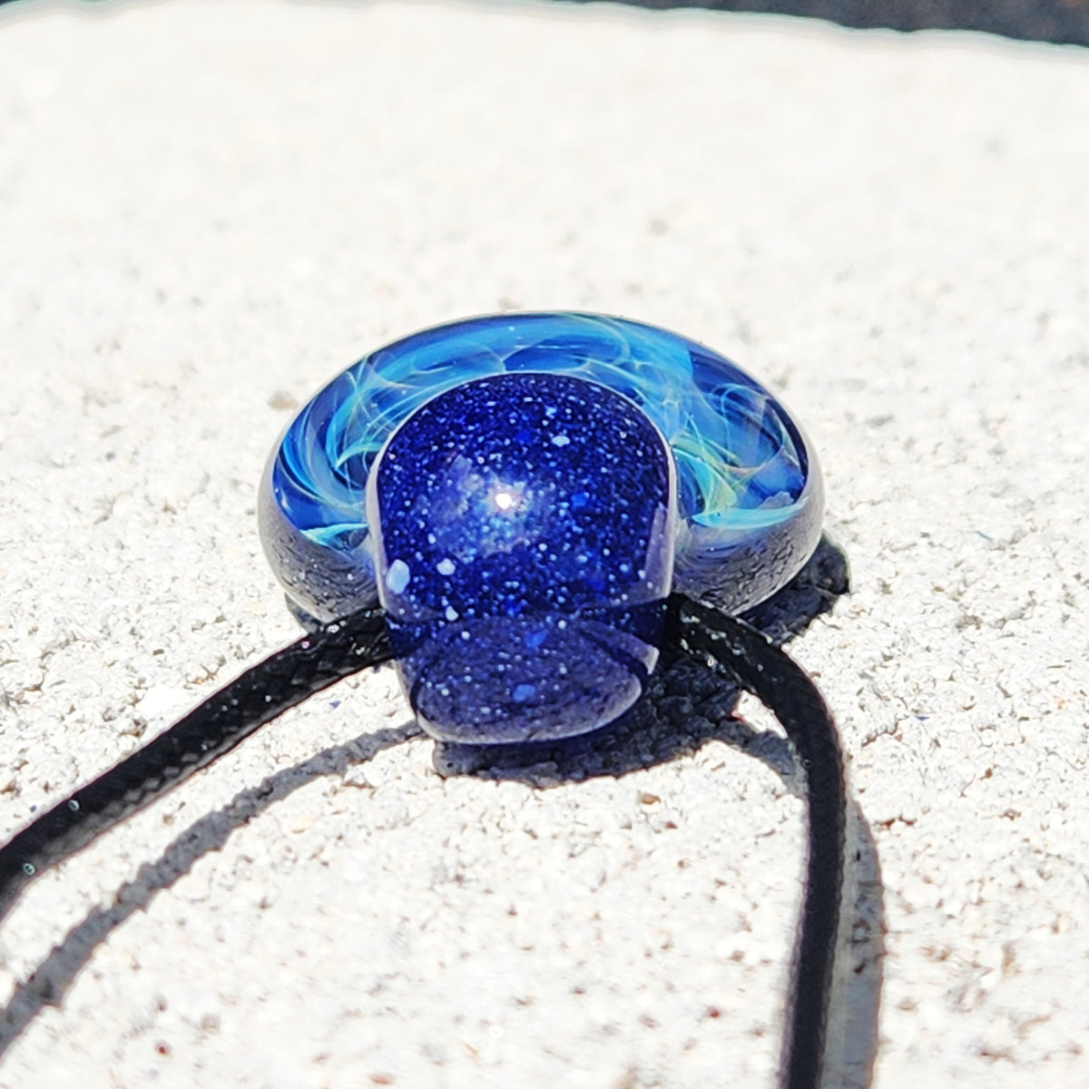 A close-up of a blue glass pendant necklace with a dark blue glittery center and lighter blue marbled swirls, threaded on a black cord over a light sandy background.