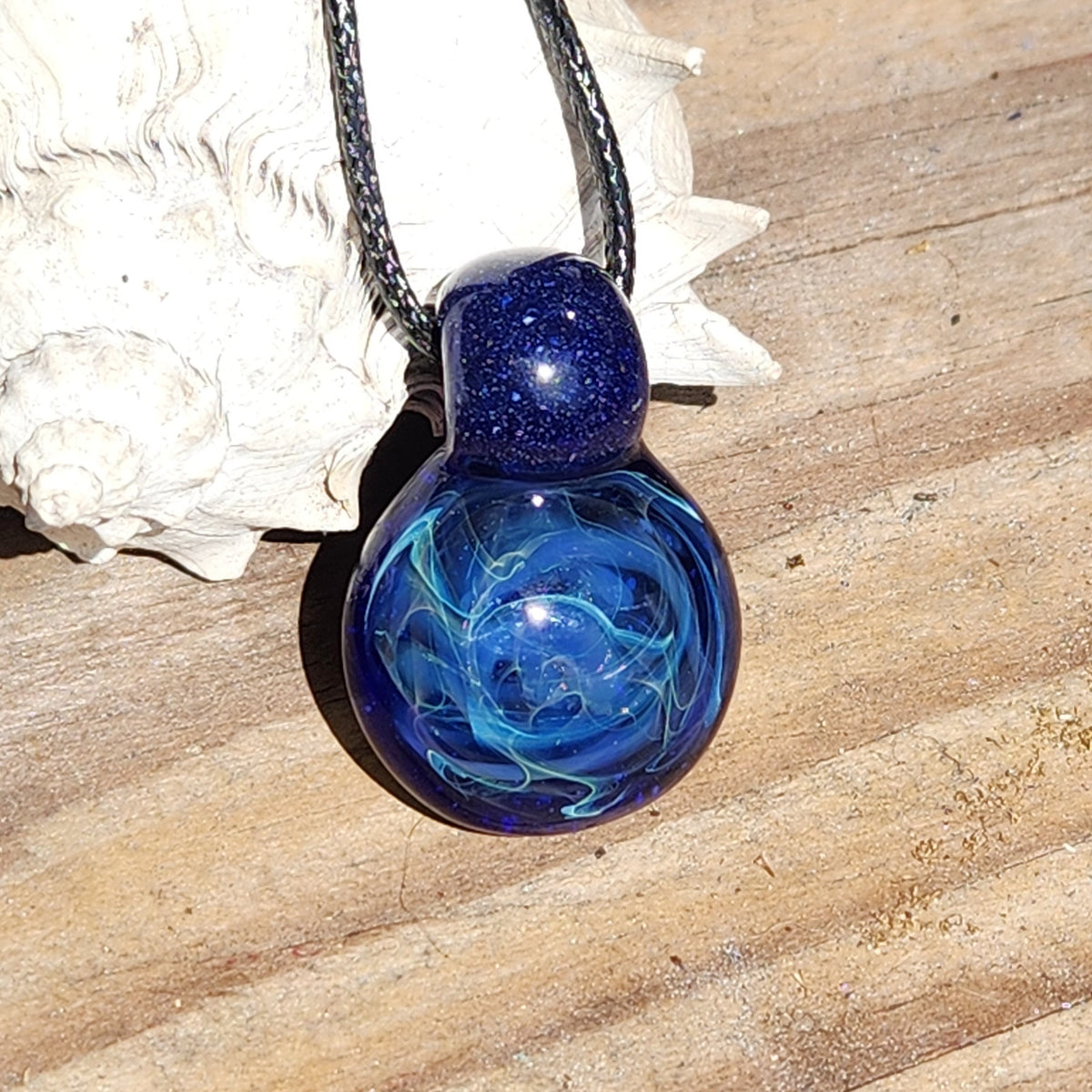 A glass pendant necklace with a deep blue and light blue marbled design, featuring a dark blue glittery top section and a black braided cord, resting on a wooden surface next to a white seashell.