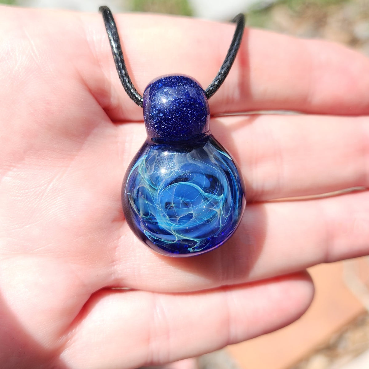 A hand holding a glass pendant necklace with a deep blue and light blue marbled design, suspended from a black cord.