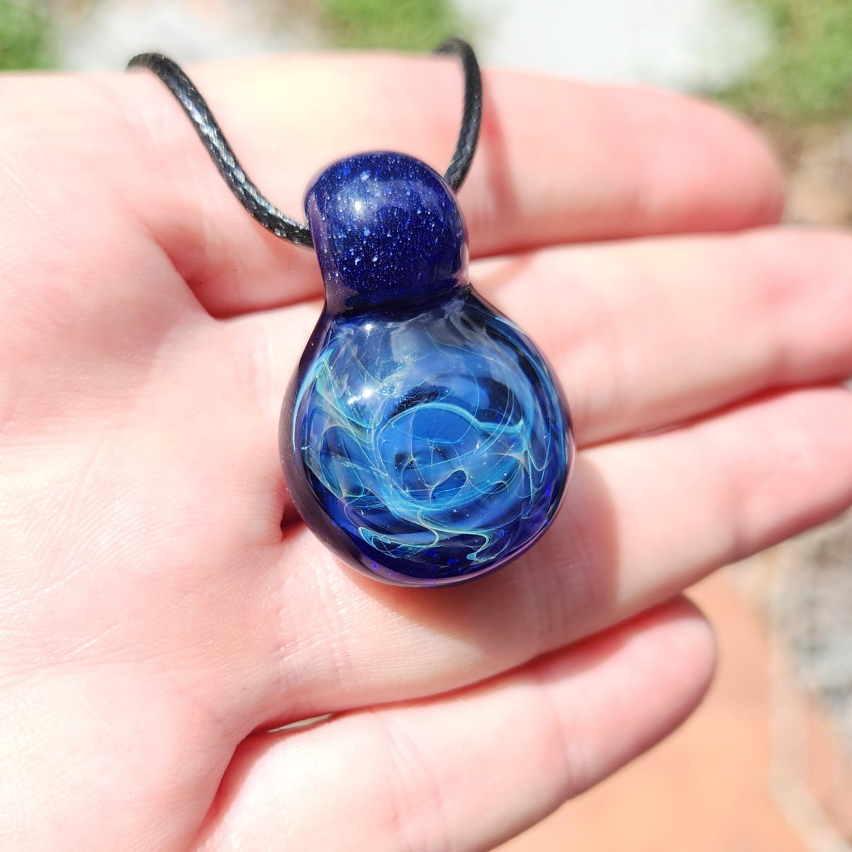 A hand holding a small glass pendant necklace with a dark blue glittery top and a swirling blue glass body on a black cord, photographed outdoors with blurred background.