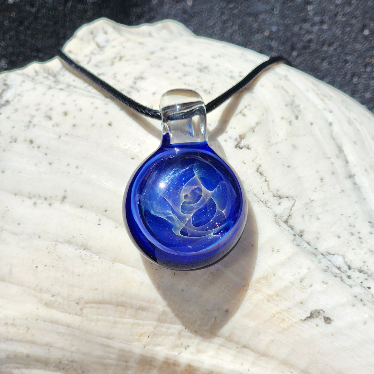 Round cobalt blue glass pendant with a pale swirling center and clear glass loop on a black cord, resting on a white seashell
