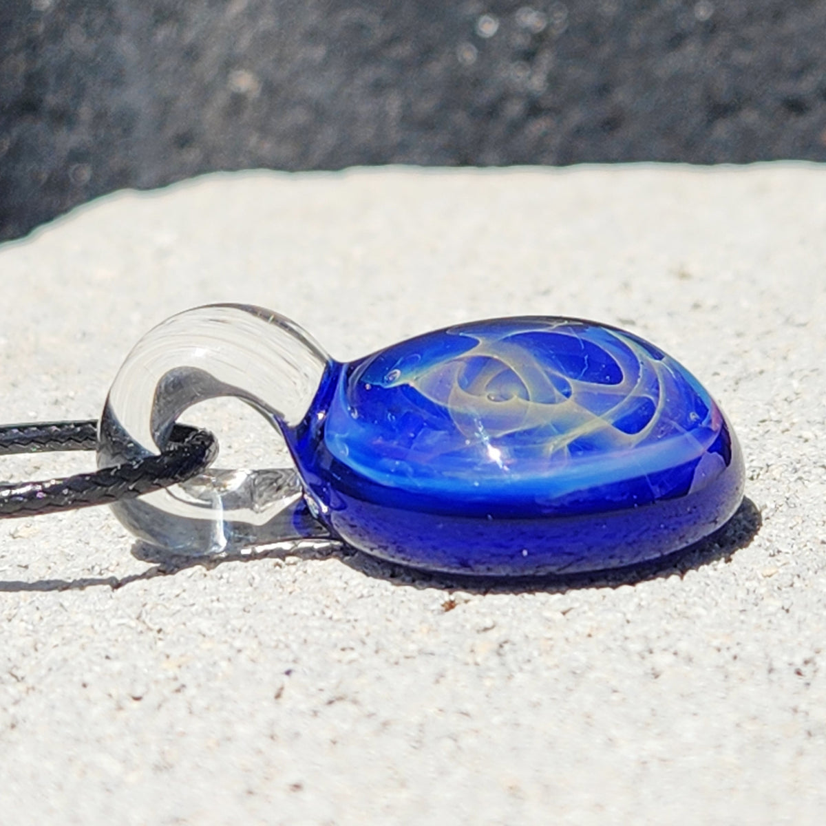 A blue and white swirled glass pendant necklace on a black cord, resting on a light concrete surface outdoors.
