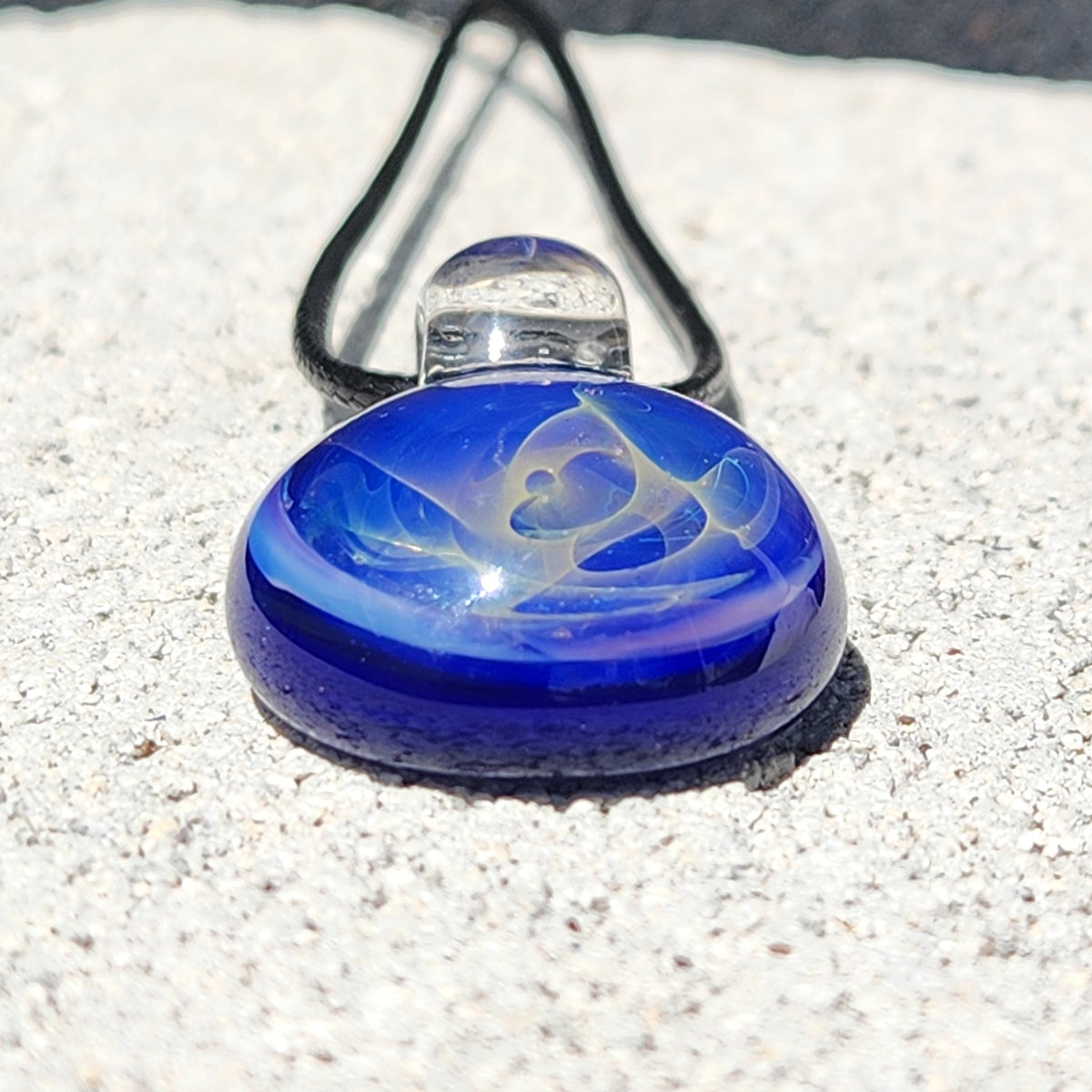 A blue glass pendant necklace with a white abstract design, resting on a light gray textured stone surface under bright sunlight.