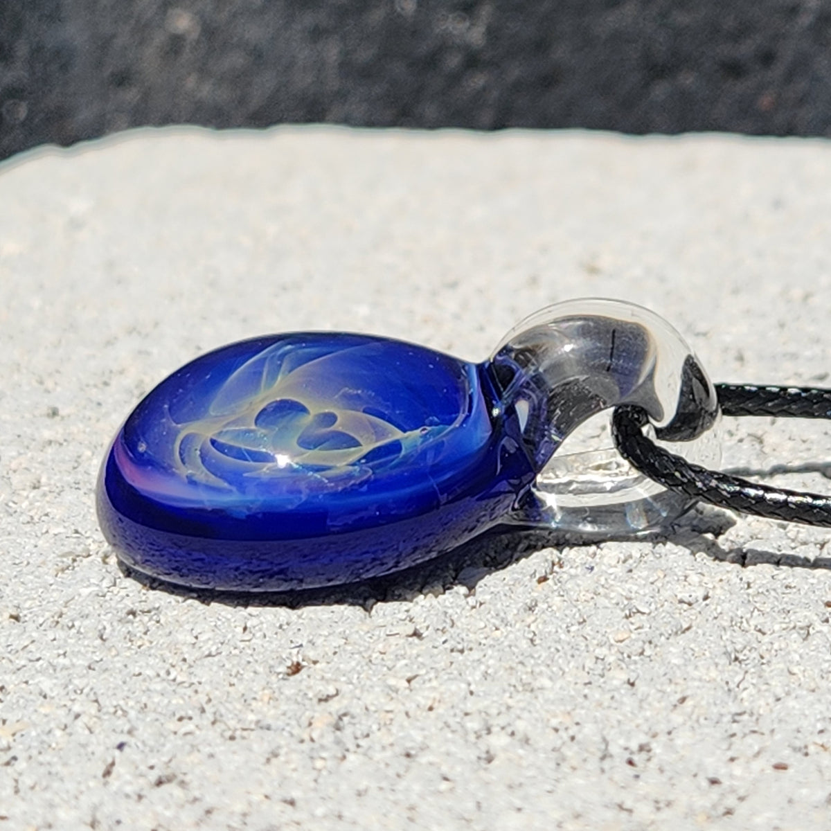 A cobalt blue glass pendant with a gold and silver swirl design, attached to a clear glass loop and black cord, resting on a light gray concrete surface.