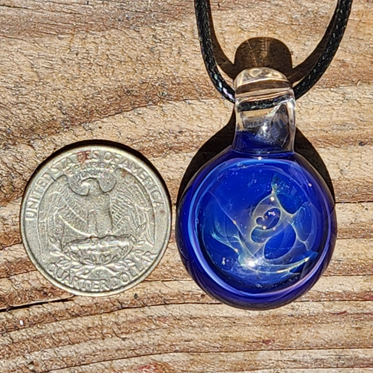 A small round blue glass pendant necklace with a black cord, shown next to a U.S. quarter coin for scale on a wooden surface.