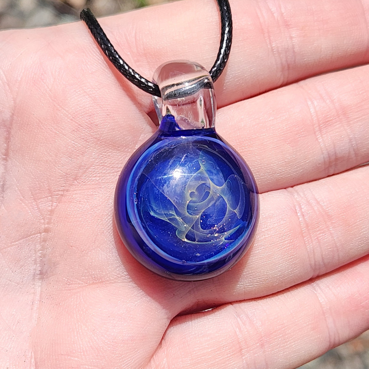 A close-up of a hand holding a round glass pendant necklace. The pendant is deep cobalt blue with a white swirling pattern inside, suspended from a black cord with a clear cap.