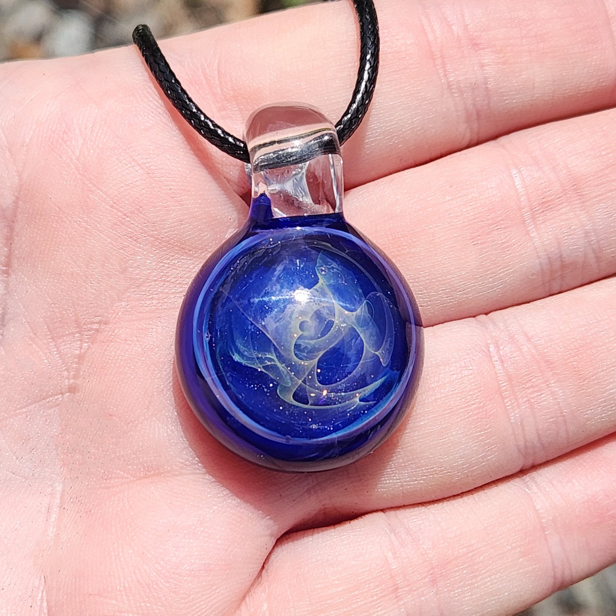 A close-up of a hand holding a round glass pendant necklace. The pendant is deep cobalt blue with swirling white and light blue patterns inside, suspended from a black cord.