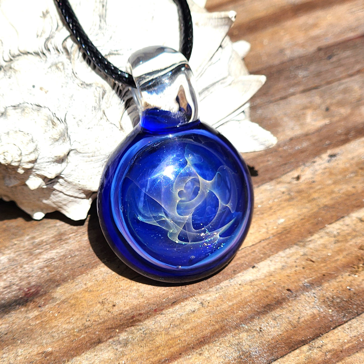 A round cobalt blue glass pendant necklace with a white floral swirl design inside, suspended from a black cord and resting on a wooden surface next to a white seashell.
