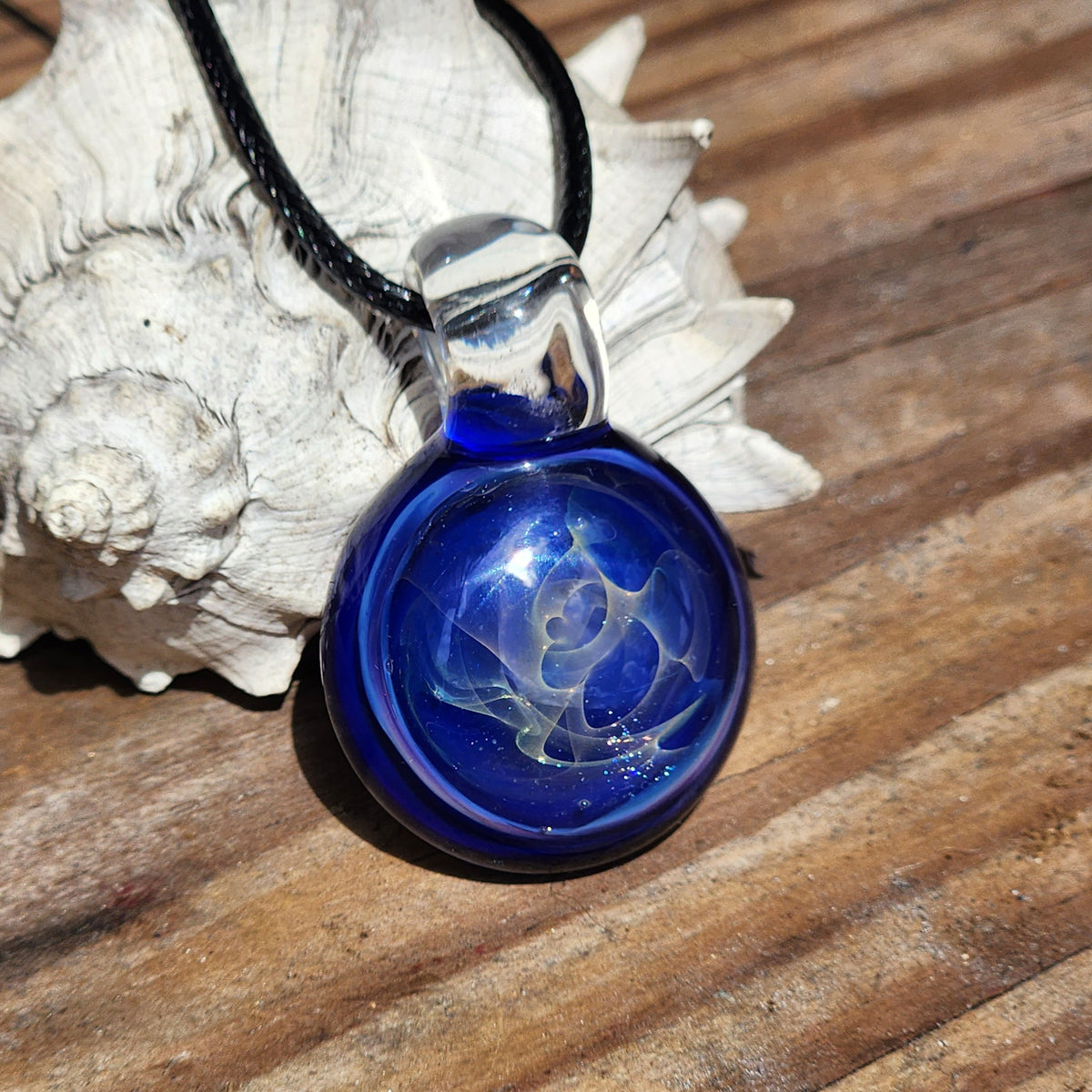 A round cobalt blue glass pendant necklace with a gold and silver fuming design inside, suspended from a black cord, resting on a white seashell over a wooden surface.