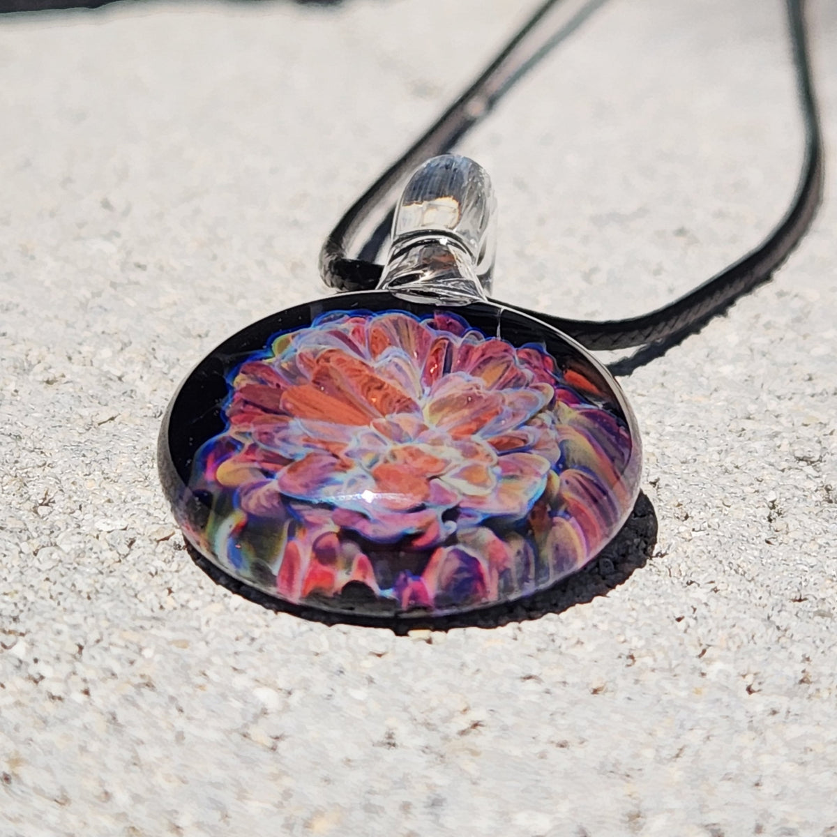 Close-up of the handmade glass pendant with vibrant floral pattern, clear glass bead, and black braided cord, on a textured surface.