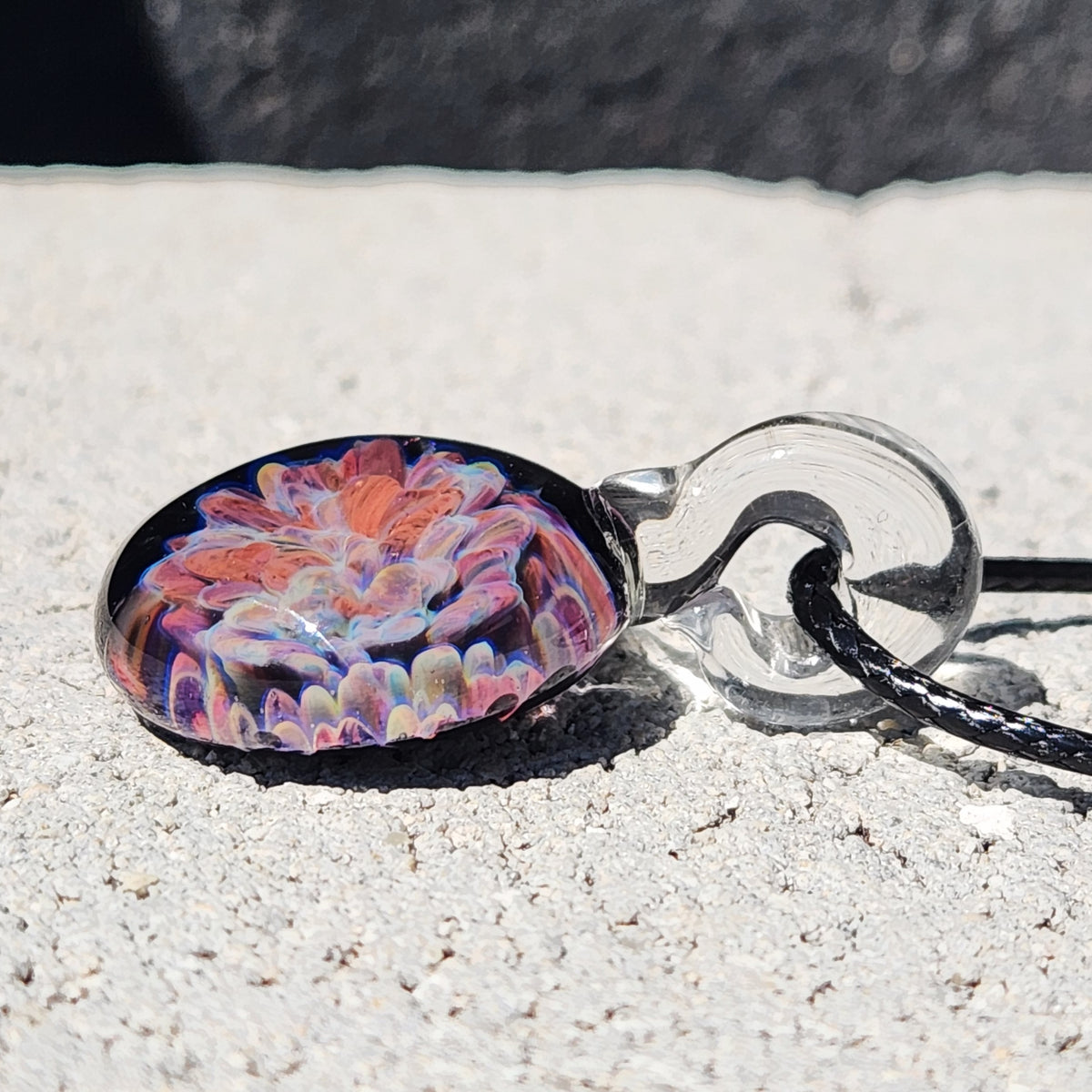 Side view of the handmade glass pendant with floral design, clear glass bead, and black braided cord, on a textured background.