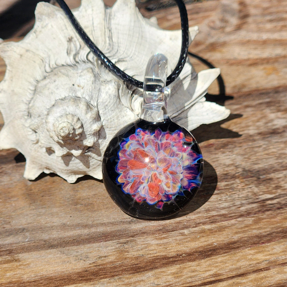 Handmade glass pendant necklace with colorful floral design, black braided cord, and clear glass bead, displayed on a textured wooden surface.