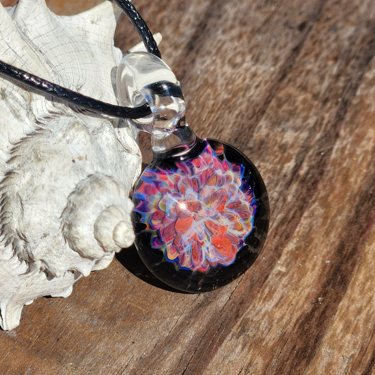 Close-up of handmade glass pendant with vibrant floral pattern, black cord, and clear glass detail, worn on finger for size reference