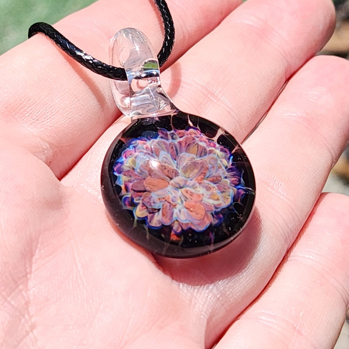 Handmade floral glass pendant necklace with black braided cord, shown on a person's hand with textured background.