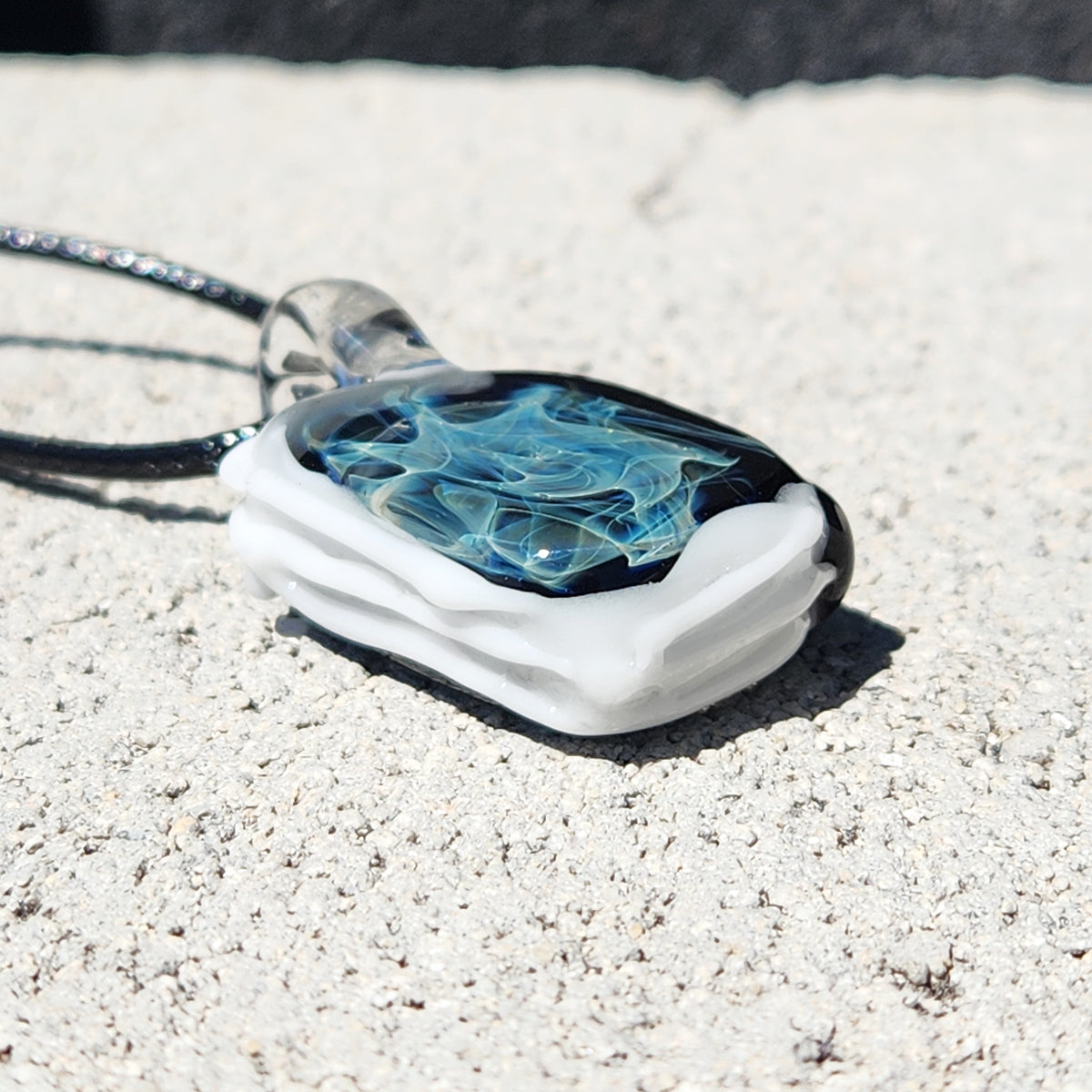 A handmade glass pendant necklace with a white layered base, a black top layer, and swirling blue and clear glass patterns, resting on a textured light gray concrete surface.