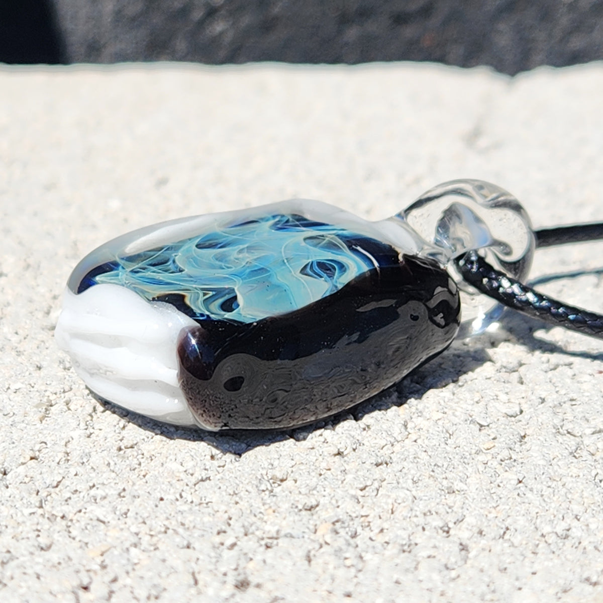 A hand-blown glass pendant necklace with a black, white, and blue marbled design, resting on a textured light gray stone surface.