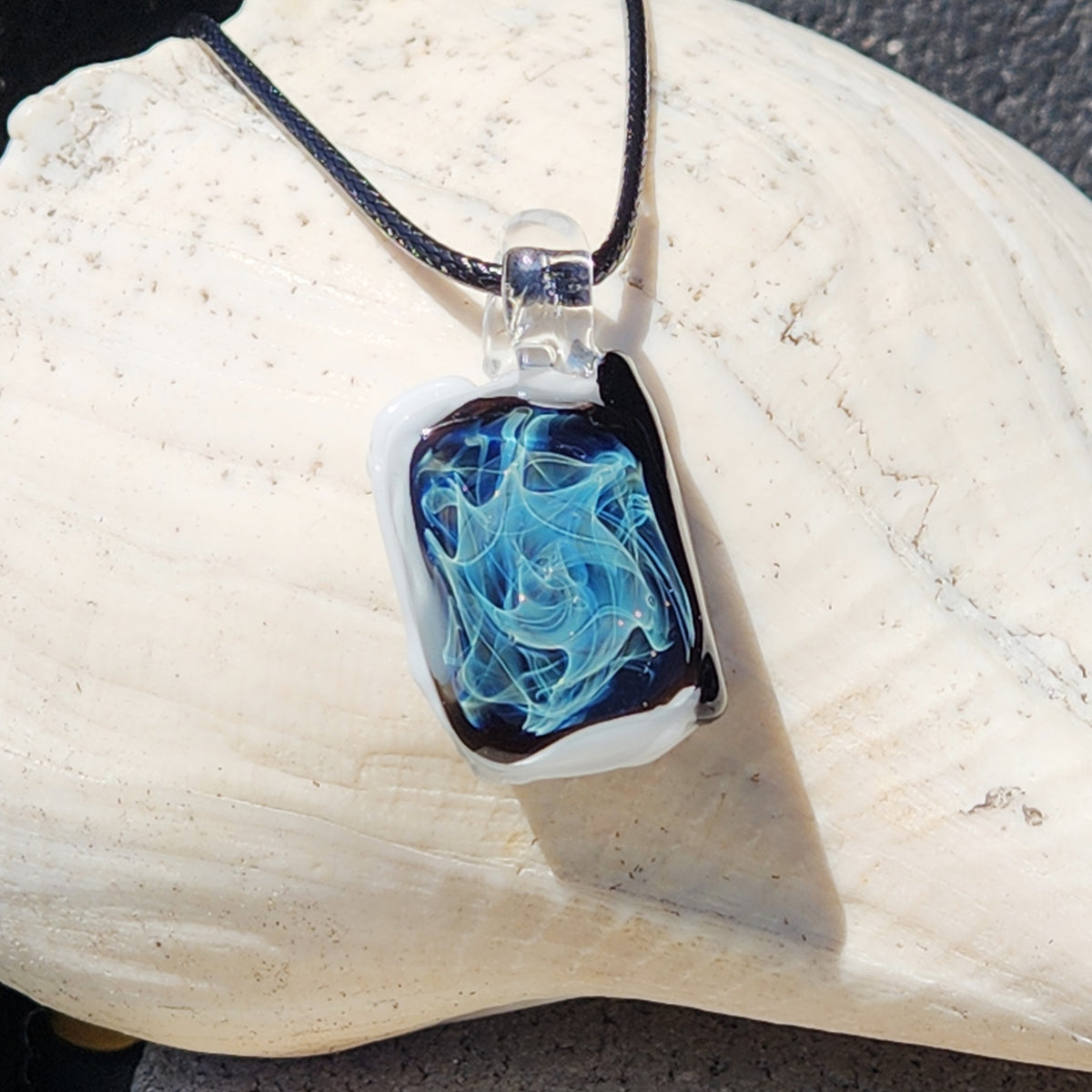 A rectangular glass pendant necklace with a black and white border and swirling blue glass pattern, hanging from a black cord on a large white seashell.