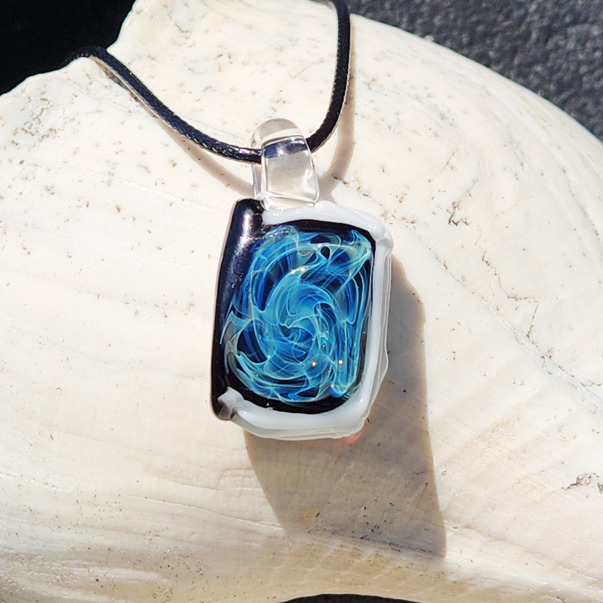 A handmade glass pendant necklace with a swirling blue and white design inside a clear glass frame, hanging from a black cord on a large white seashell.