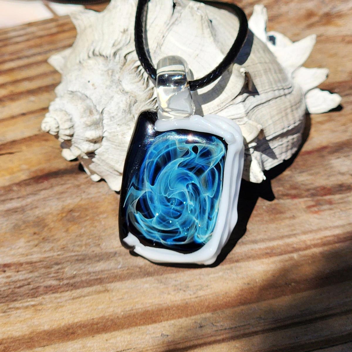 A rectangular glass pendant necklace with a black cord, featuring a swirling blue and turquoise design over a black and white layered glass background, displayed on a wooden surface with a seashell prop.
