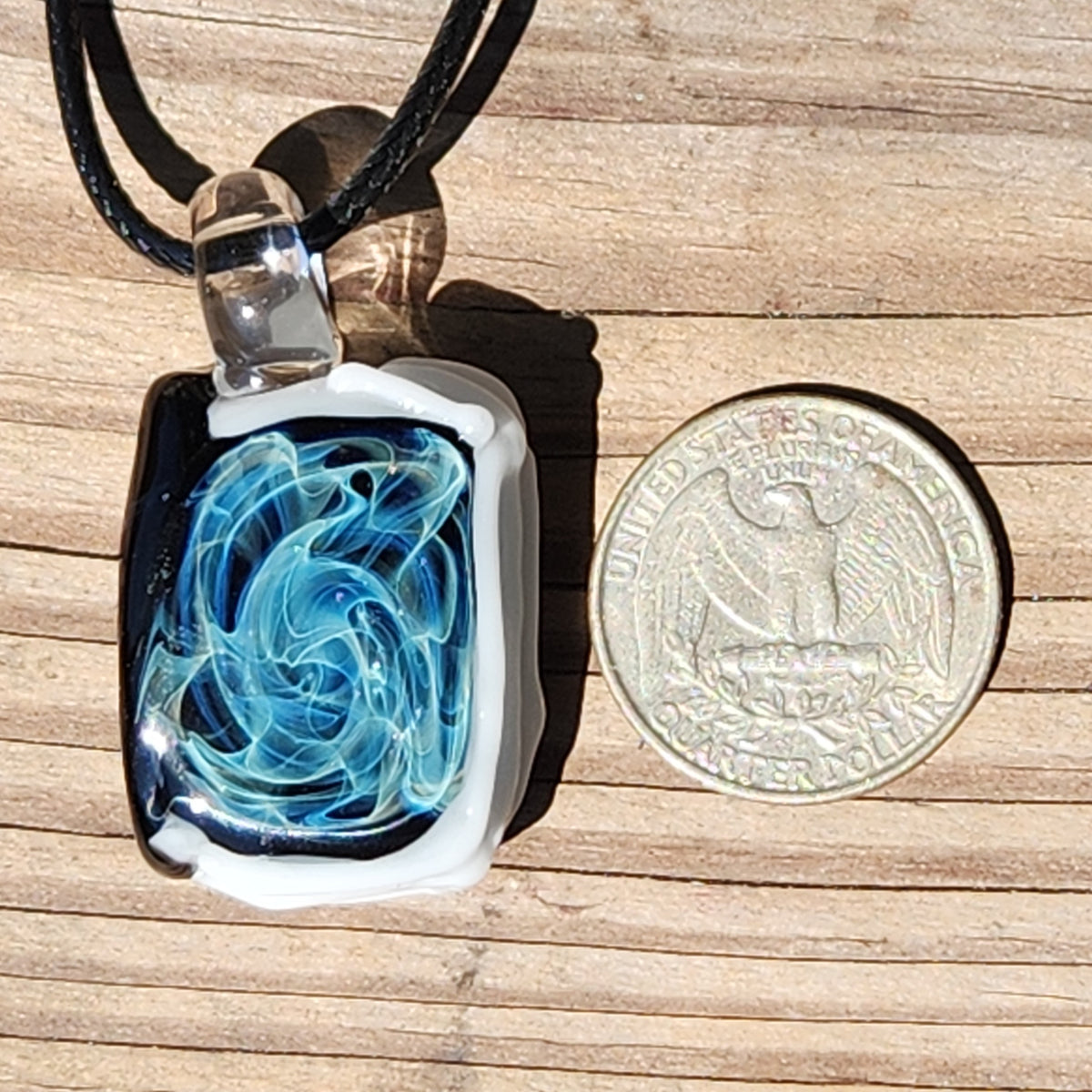 A rectangular glass pendant necklace with a swirling blue and white design inside a clear glass frame, resting on a wooden surface next to a US quarter coin for scale. The pendant is suspended from a black cord.