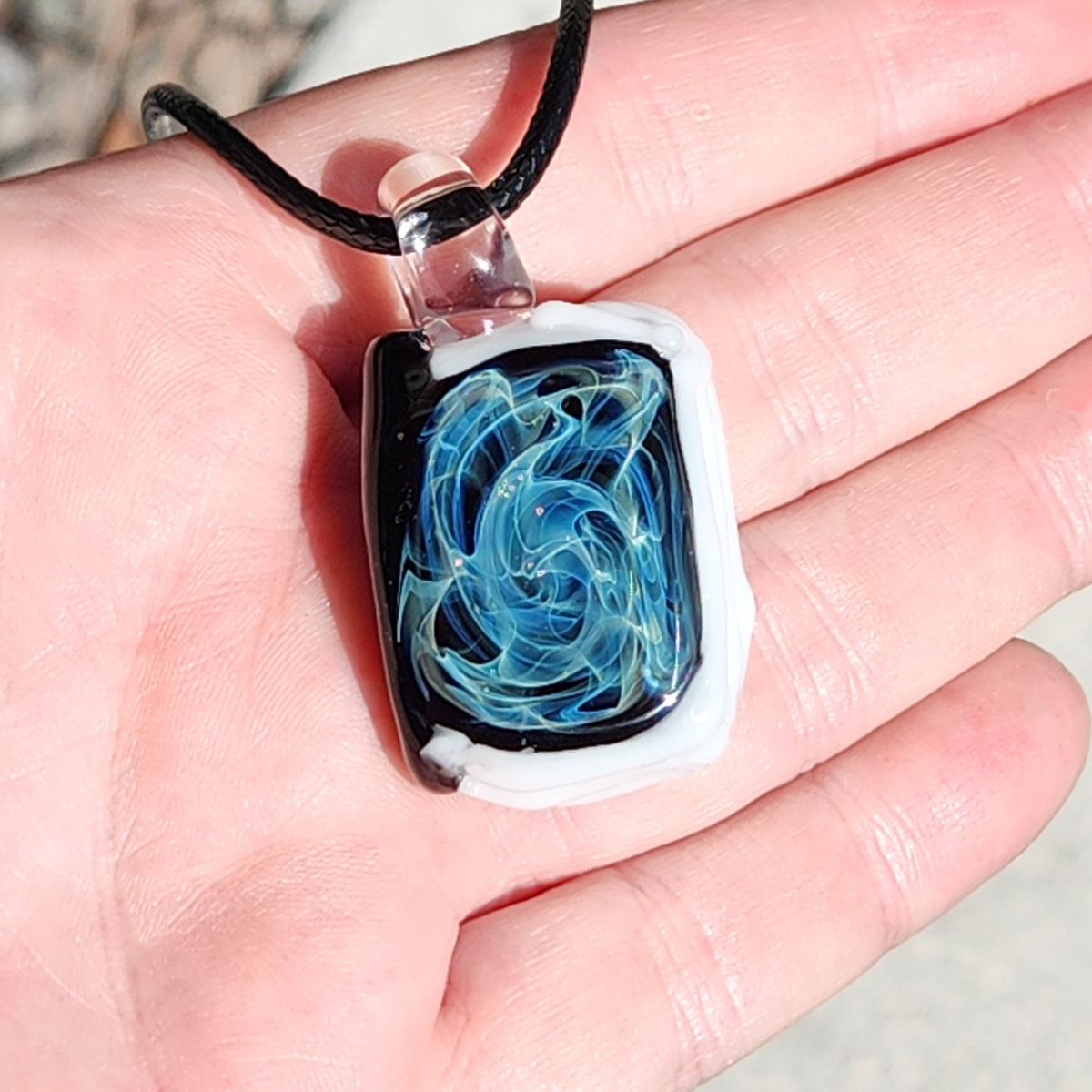 A rectangular glass pendant necklace with a black and white border and a swirling blue and light blue design in the center, shown resting on a person's palm.