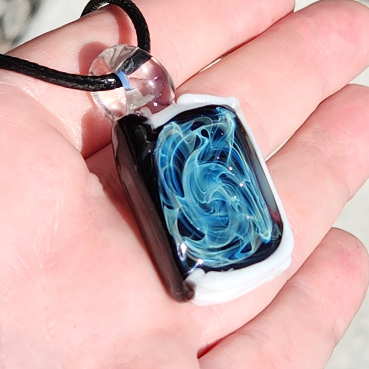 A hand holding a rectangular glass pendant necklace with a black cord. The pendant features a black and white border with a swirling blue and light blue design inside, and a clear glass bead at the top.