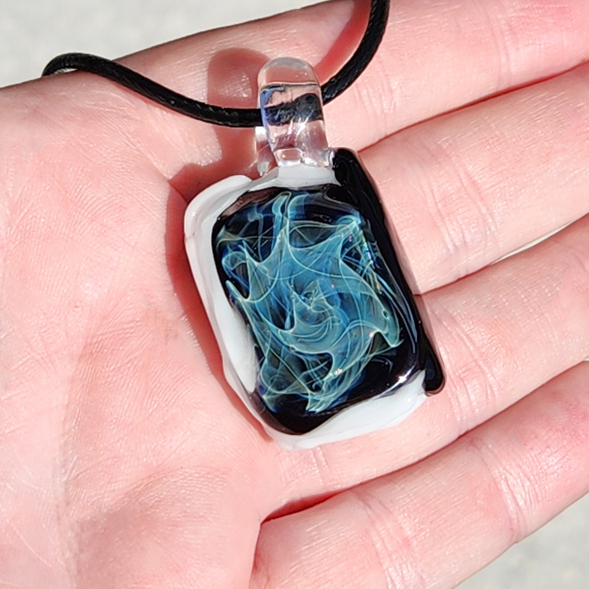 A hand holding a square glass pendant necklace with a black cord. The pendant features a white border and a swirling blue and black glass design in the center.