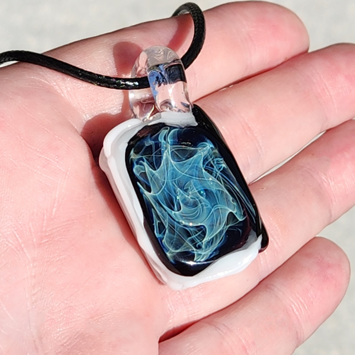 A hand holding a rectangular glass pendant necklace with a black cord. The pendant features a smoky, marbled design with swirls of blue, black, and white, set in a clear glass frame.