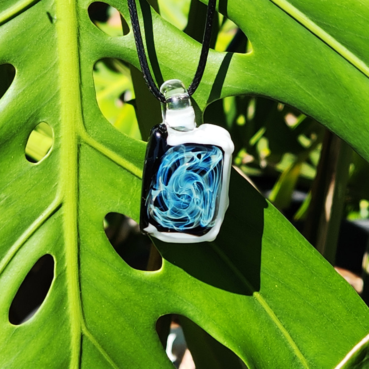 A rectangular glass pendant necklace with a black and white border and a swirling blue and white design inside, hanging from a black cord against a large green monstera leaf.