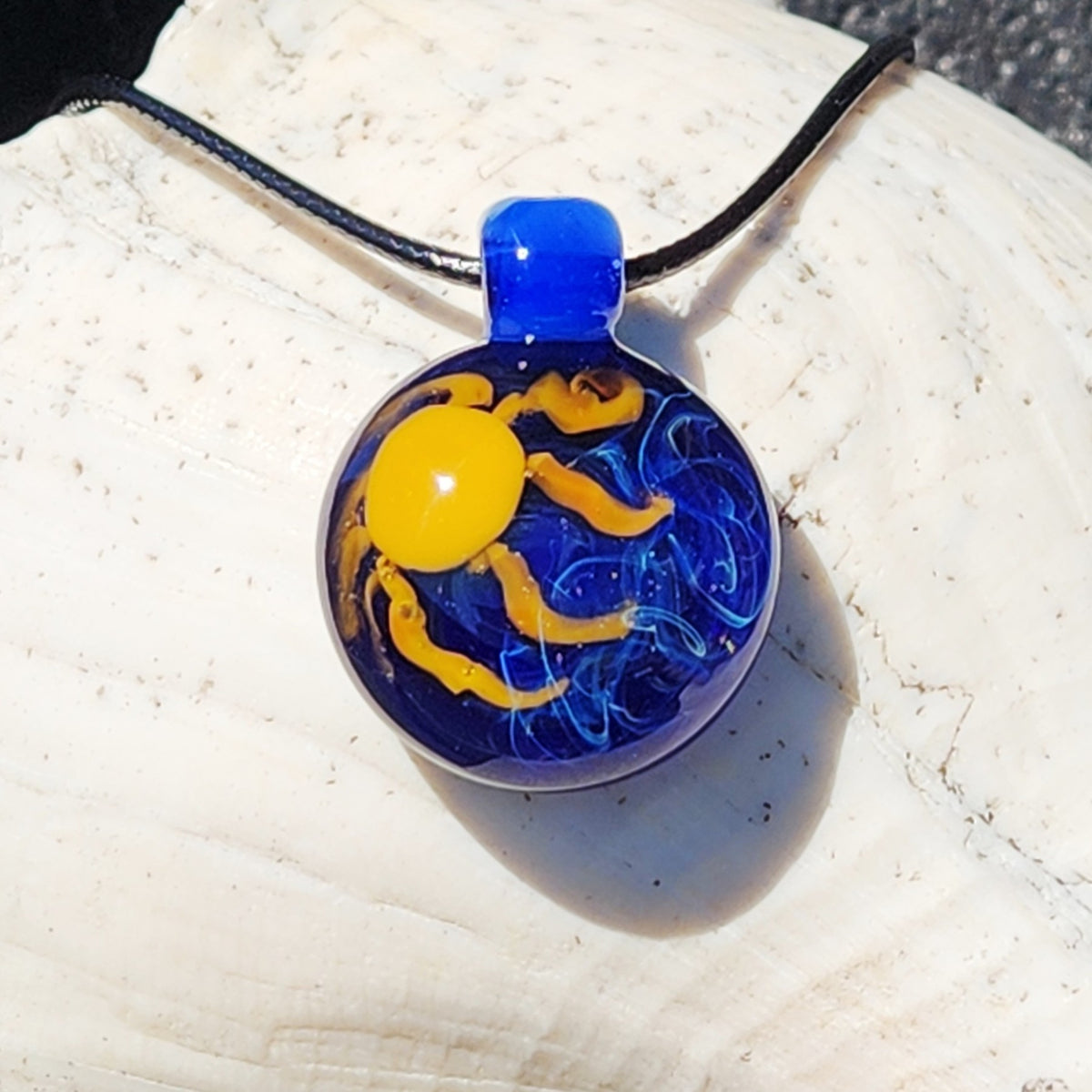 A round glass pendant necklace featuring a yellow sun and blue wavy lines on a black cord, displayed on a white seashell.