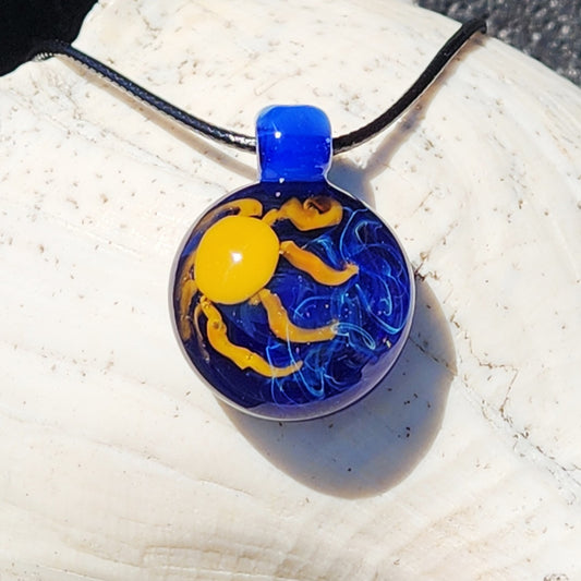 A round glass pendant necklace featuring a yellow sun and blue wavy lines on a black cord, displayed on a white seashell.