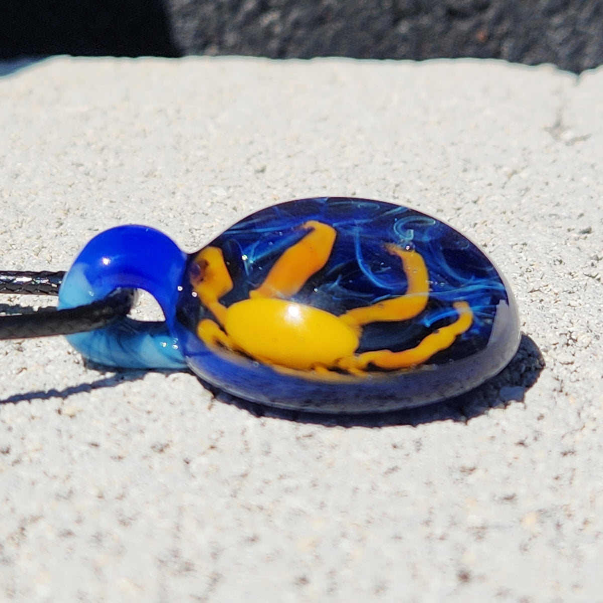 A handmade glass pendant featuring a blue and yellow design, resting on a light-colored textured surface.