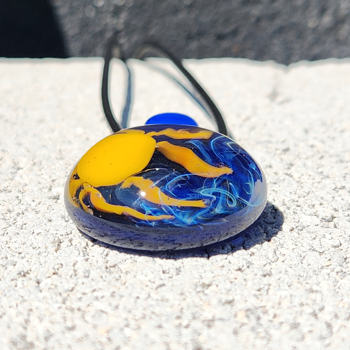 A hand-blown glass pendant necklace with a blue and yellow design, resting on a textured light gray surface with a dark background.