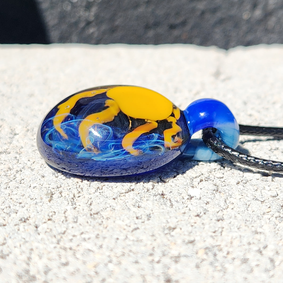 A handmade glass pendant necklace with a blue and yellow design, hanging from a black cord against a textured light gray concrete wall.