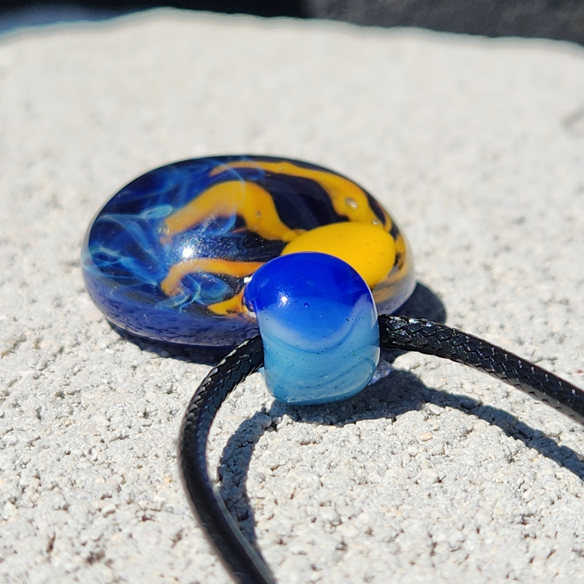 A close-up of a glass pendant necklace with a marbled blue, yellow, and black design, threaded on a black cord against a textured light gray concrete background.