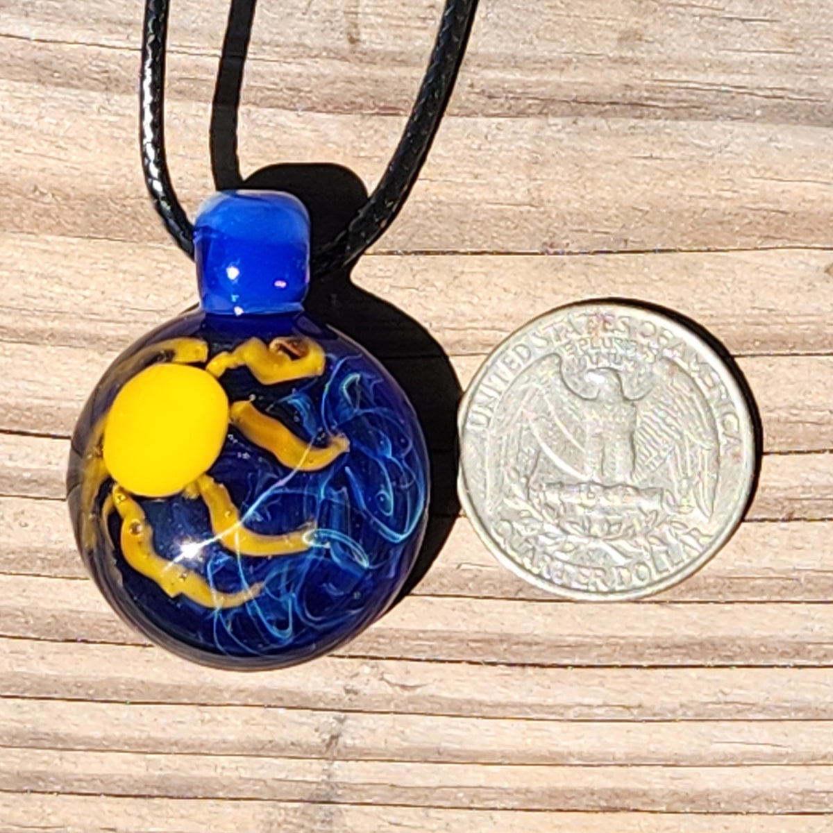 A small round glass pendant necklace with a black cord, featuring a blue and yellow design, shown next to a US quarter coin on a wooden surface.