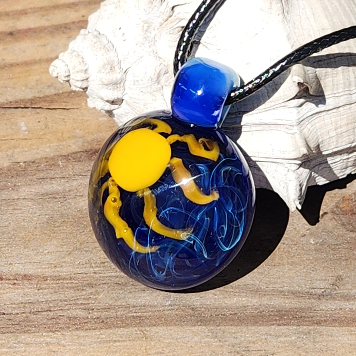 A handmade glass pendant necklace featuring a blue and yellow design inside a clear glass sphere, suspended from a black cord and resting on a white seashell over a wooden surface.