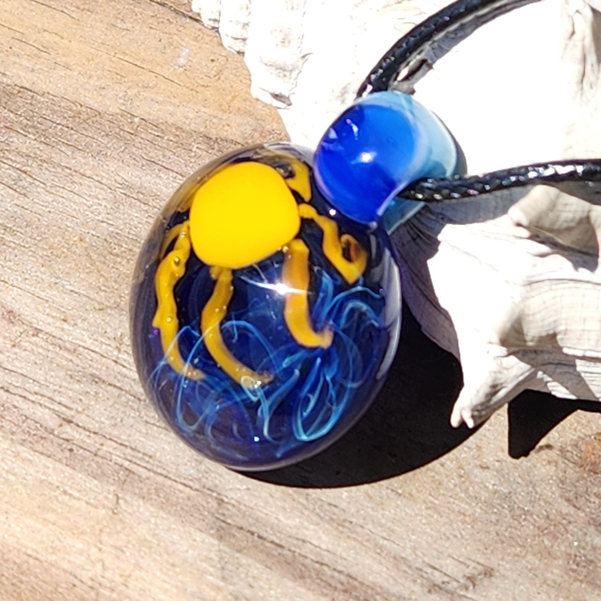 A handmade glass pendant necklace featuring a blue and yellow octopus design inside a clear glass sphere, resting on a white seashell over a wooden surface.