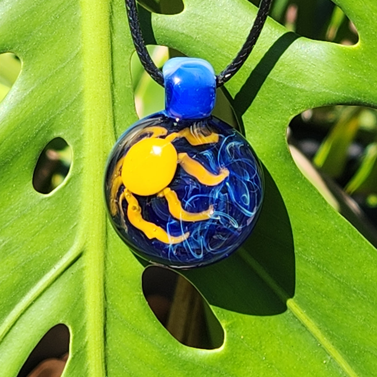 A round glass pendant necklace featuring a yellow sun and blue wavy patterns, suspended on a black cord and resting on a large green leaf.
