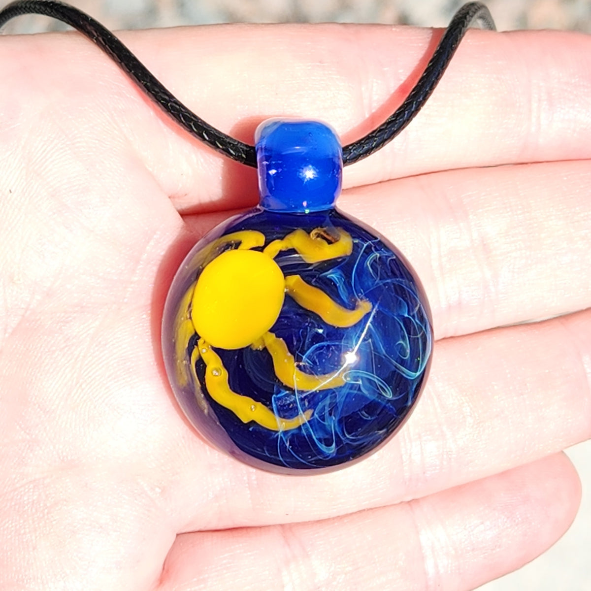 A hand holding a small round glass pendant necklace with a blue and yellow sun design on a black cord.