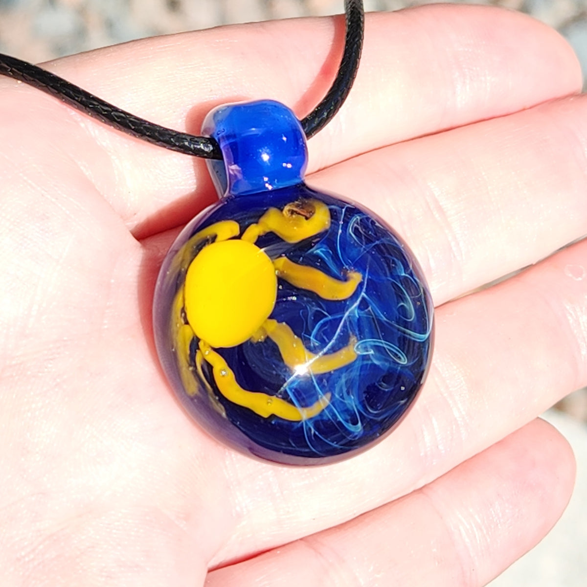 A hand holding a round glass pendant necklace with a deep blue background, a bright yellow sun motif, and light blue wavy details, suspended from a black cord.