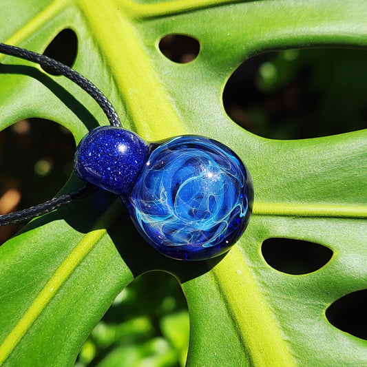 A blue glass pendant necklace with a black cord, featuring a smaller round blue bead and a larger swirling blue glass sphere, resting on a green monstera leaf.