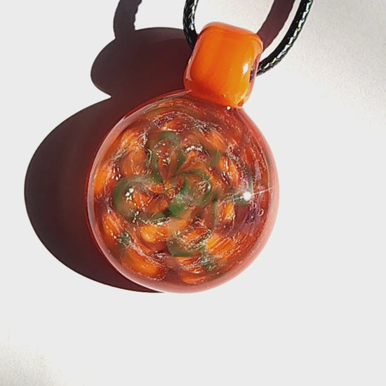Sunlit view of handmade orange swirl glass pendant necklace with shimmering silver and green accents on a white background.