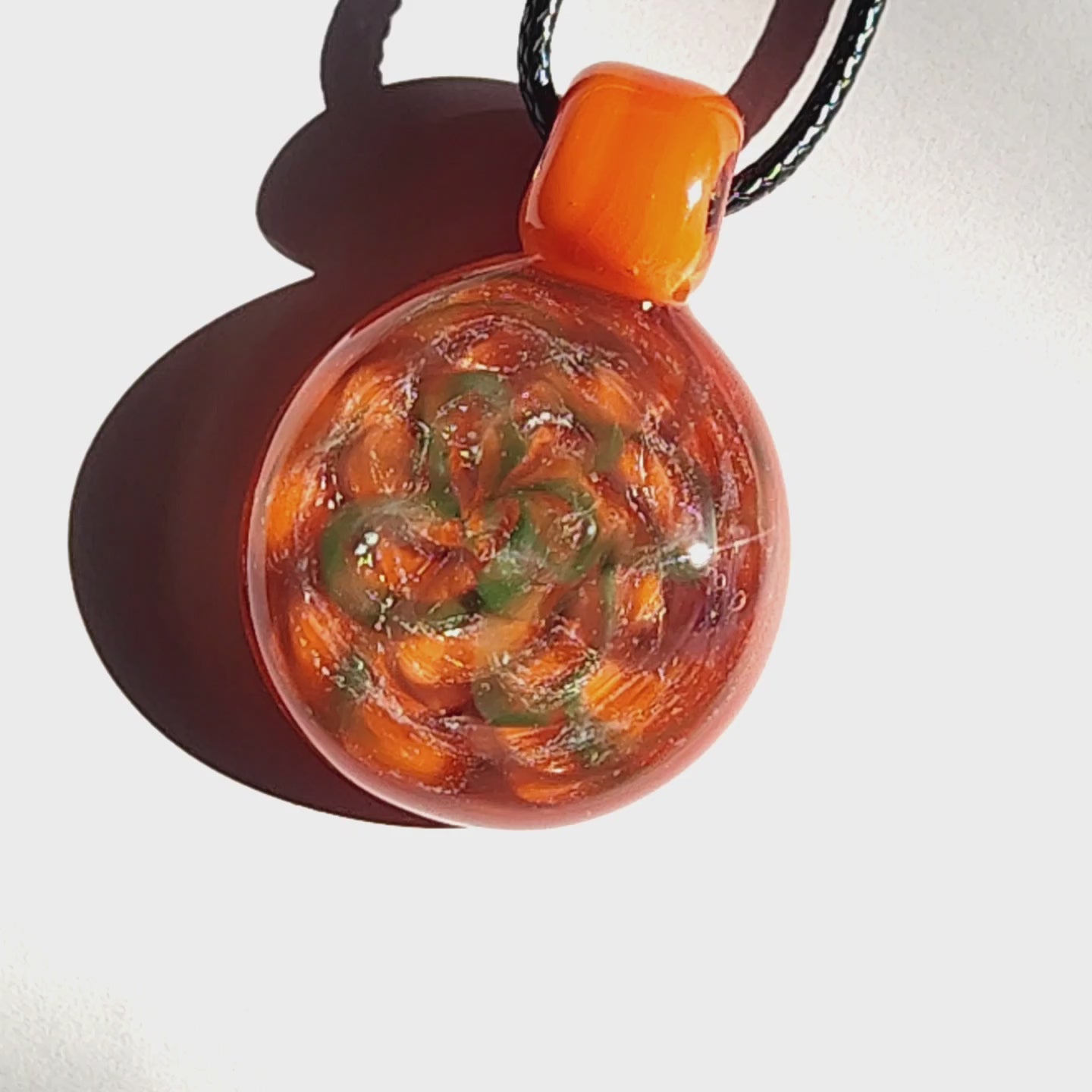 Sunlit view of handmade orange swirl glass pendant necklace with shimmering silver and green accents on a white background.