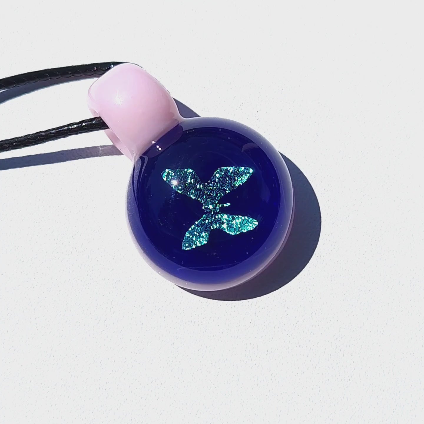 Blue glass butterfly pendant with pink loop and black cord, shown against a white background for detailed product view.