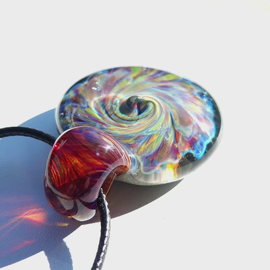Close-up of multicolor glass pendant necklace showing iridescent swirl and red-amber loop, handmade artisan jewelry.